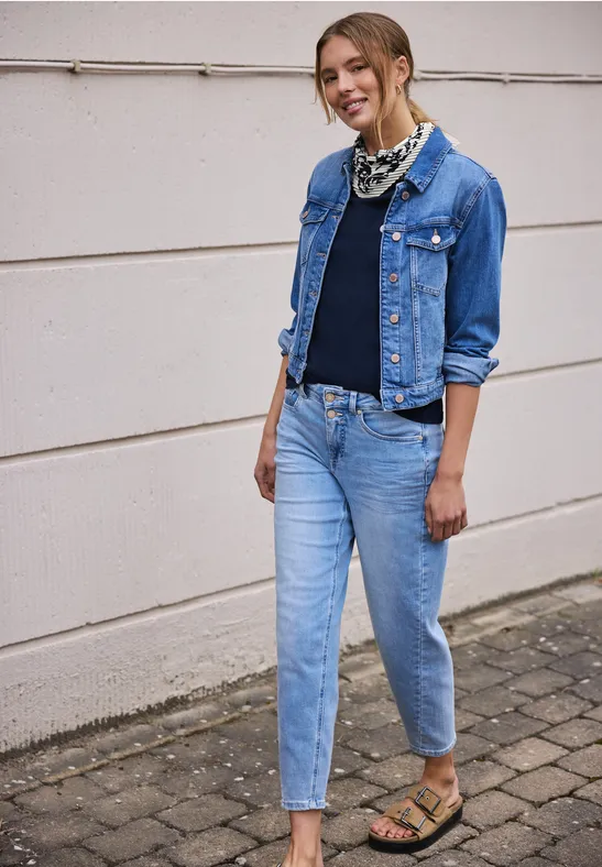 Person in Jeansjacke, Jeanshose, schwarzem Shirt, Sandalen und Halstuch steht vor heller Wand.