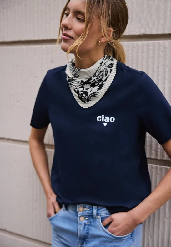 Frau in dunkelblauem T-Shirt mit 'ciao ♥'-Aufdruck, gestreiftem Halstuch und Jeans, vor Hauswand stehend