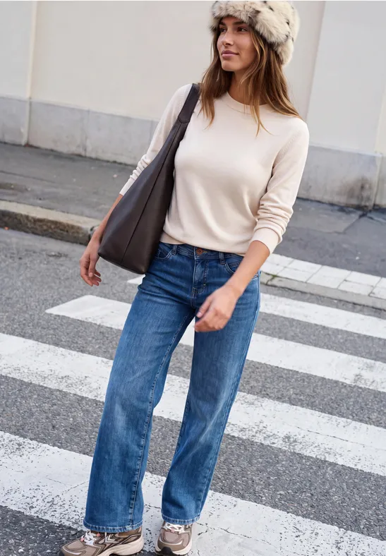 Eine Frau in weitem Jeans, hellrosa Pullover und Fellmütze überquert einen Zebrastreifen mit Tasche.