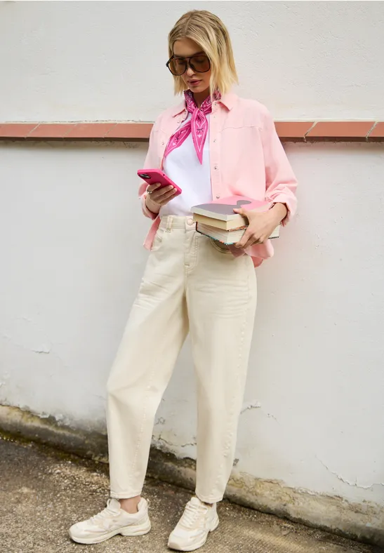 Eine junge Frau in rosa Bluse lehnt an einer Wand, hält Smartphone und Bücher in der Hand.