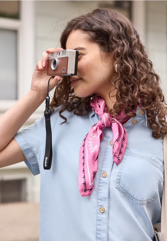 Personne aux cheveux bouclés en chemise denim et foulard rose prenant une photo avec un appareil.