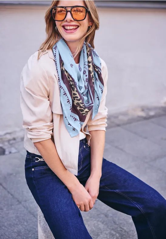 Eine junge Frau mit Sonnenbrille, Halstuch und Jeans lehnt lächelnd an einer Hauswand.