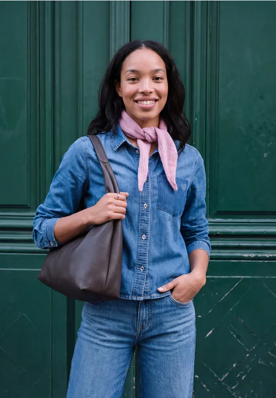 Vrouw in denim overhemd en jeans glimlachend voor een donkergroene deur, met sjaal en tas.