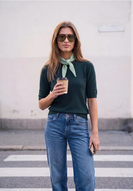 Eine Frau mit Sonnenbrille, grünem Tuch um den Hals, Pullover und Jeans hält einen Kaffee und ein Handy.