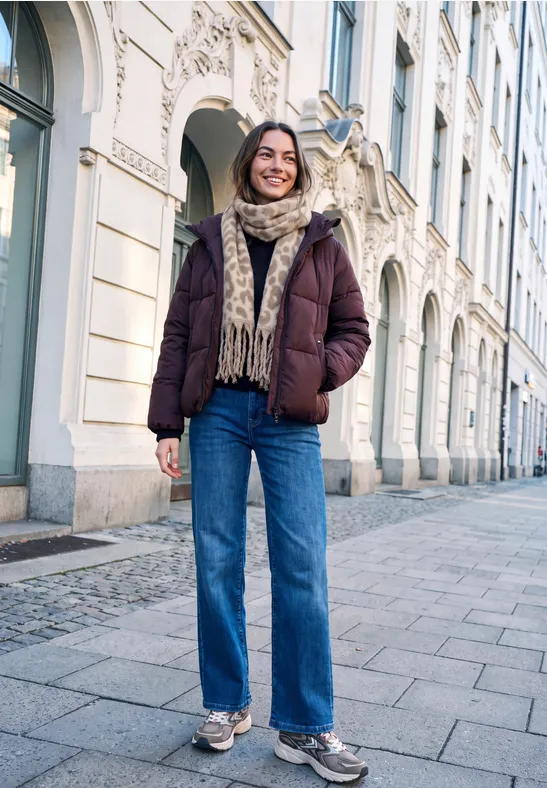 Frau in Winterjacke und Jeans geht lächelnd eine Straße mit historischen Gebäuden entlang.