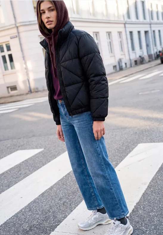 Jugendliche:r steht mit Kapuzenjacke, Jeans und Sneakern auf einem Zebrastreifen in urbaner Straße.