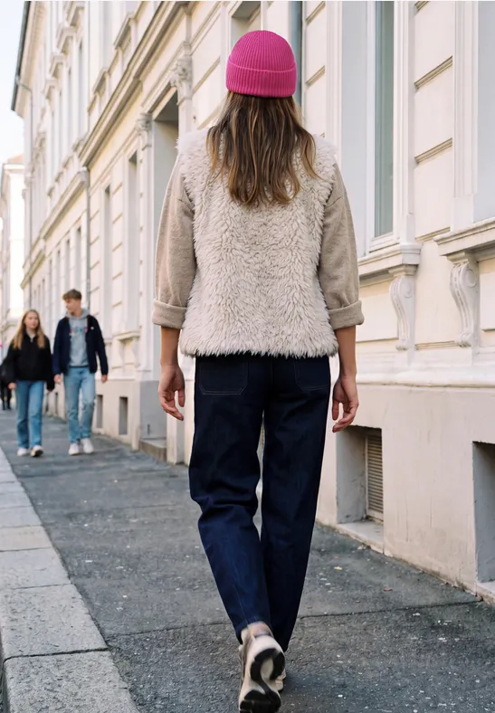 Frau mit pinker Mütze und Jeans läuft eine gepflasterte Straße entlang, im Hintergrund zwei Passanten.