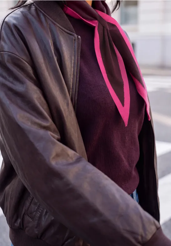 Nahaufnahme einer Person in schwarzer Lederjacke, weinrotem Pullover und pinkem Halstuch auf der Straße.
