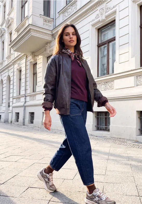 Frau in Bomberjacke, Jeans und Sneakern spaziert selbstbewusst an eleganten Stadthäusern vorbei.