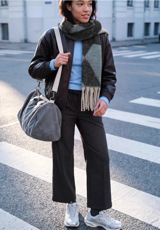 Femme en veste en cuir, écharpe et baskets blanches traversant la rue avec un sac.