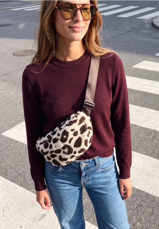 Frau mit Sonnenbrille, dunkelrotem Pullover und Jeans, trägt eine Leoparden-Bauchtasche am Zebrastreifen.