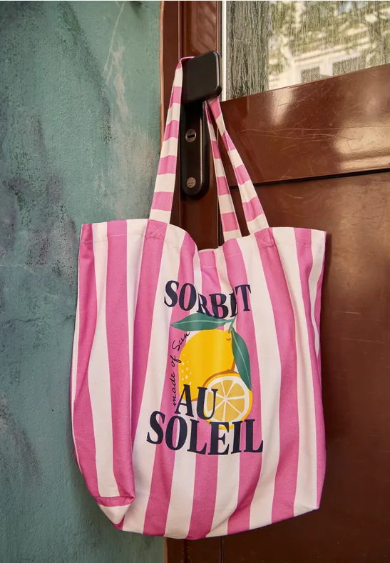 Pink-weiß gestreifte Tragetasche mit 'Sorbet au Soleil'-Aufdruck und Zitronenscheibe, an Türhaken hängend.