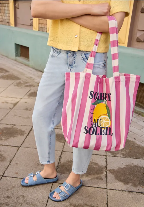 Person in Jeans und blauen Sandalen hält gestreifte Tasche mit Zitronengrafik und „Sorbet au Soleil“-Aufdruck.
