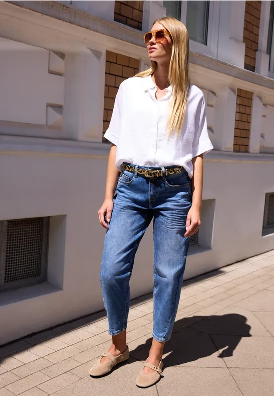 Junge Frau in weißem Hemd, blauen Jeans und Sonnenbrille lehnt entspannt an einer hellen Hausfassade.