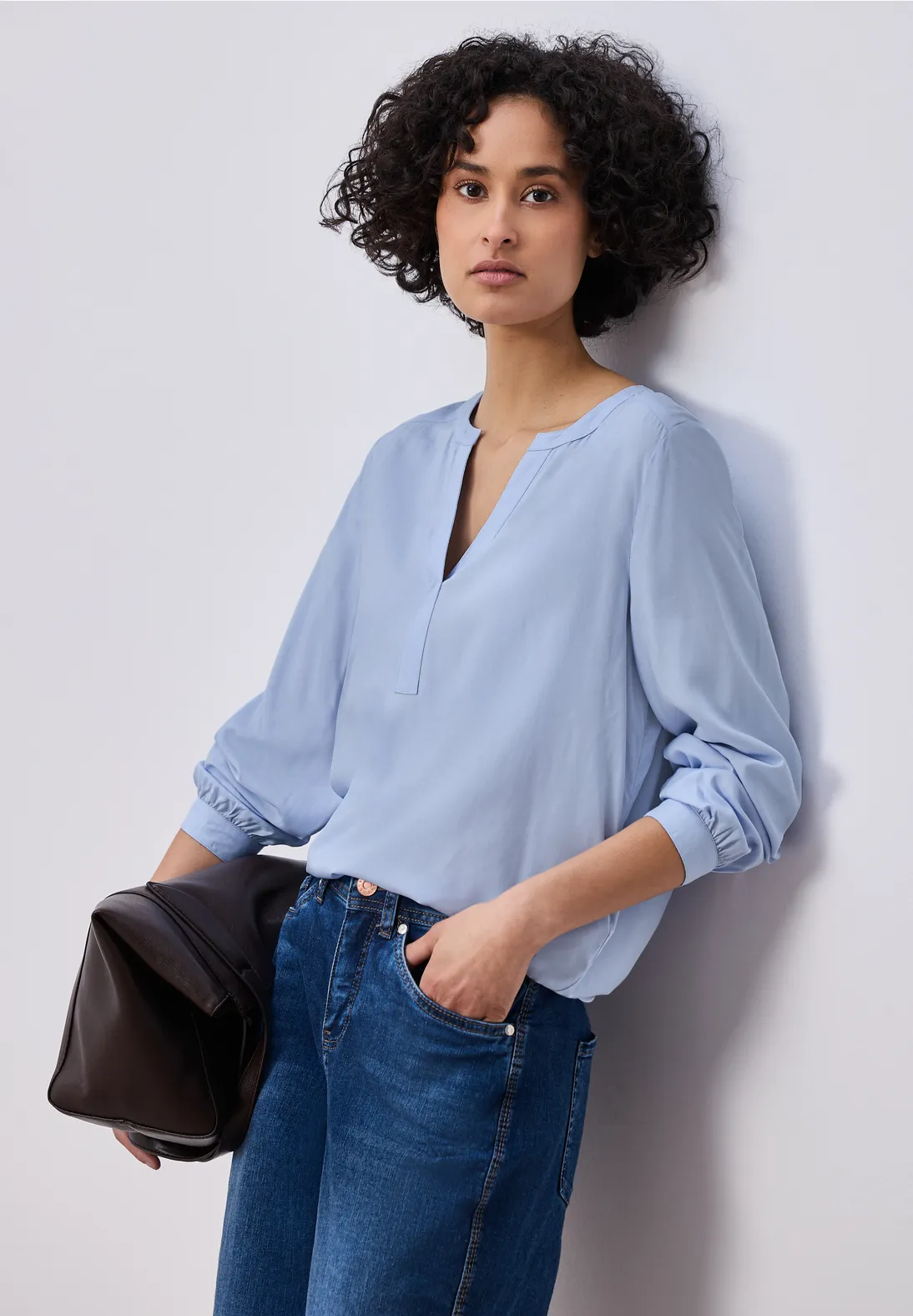 Frau in hellblauer V-Ausschnitt-Bluse mit Jeans hält schwarze Clutch vor hellem Hintergrund.
