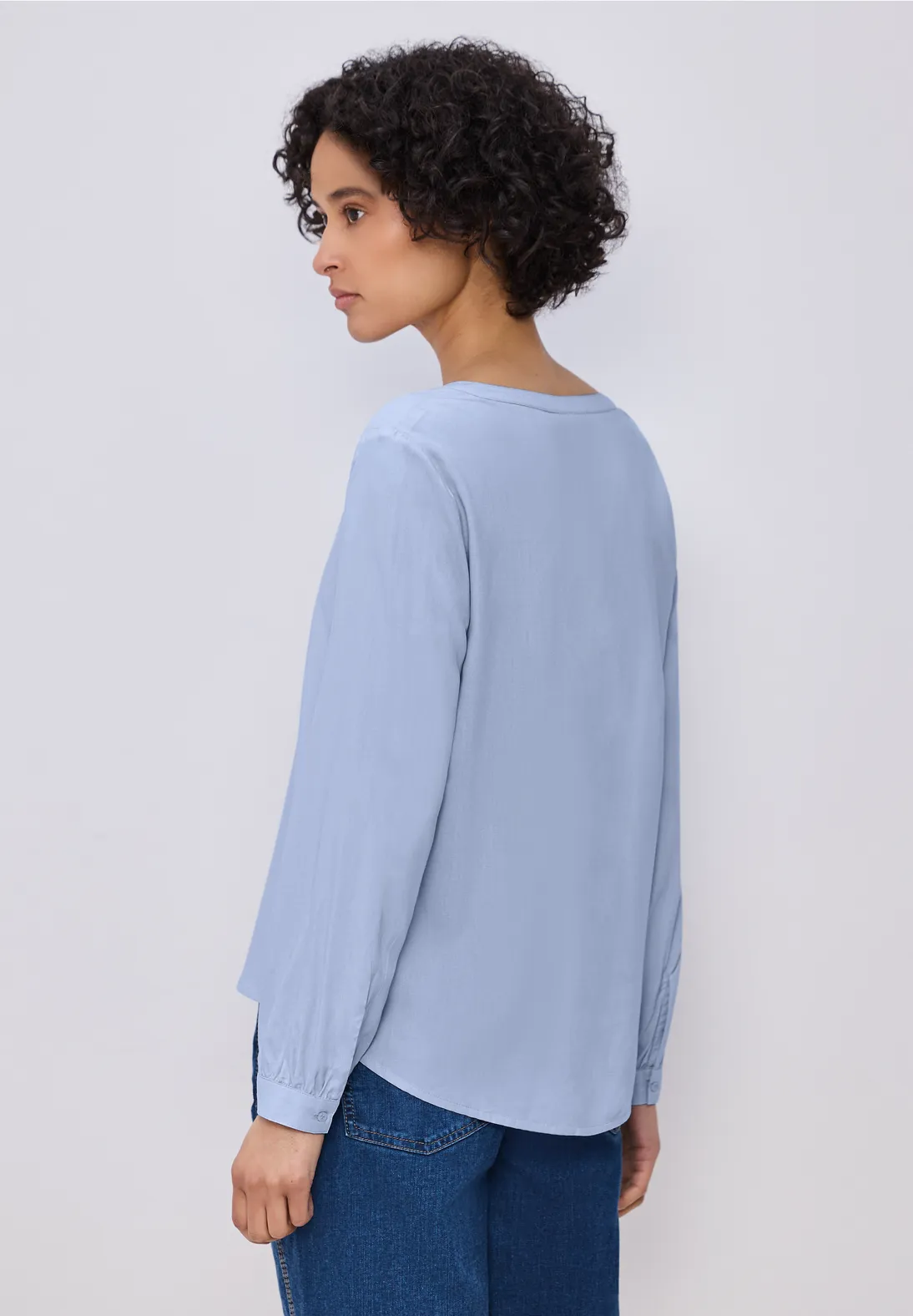 Person von hinten in hellblauer Bluse und dunklen Jeans vor neutralem Hintergrund.
