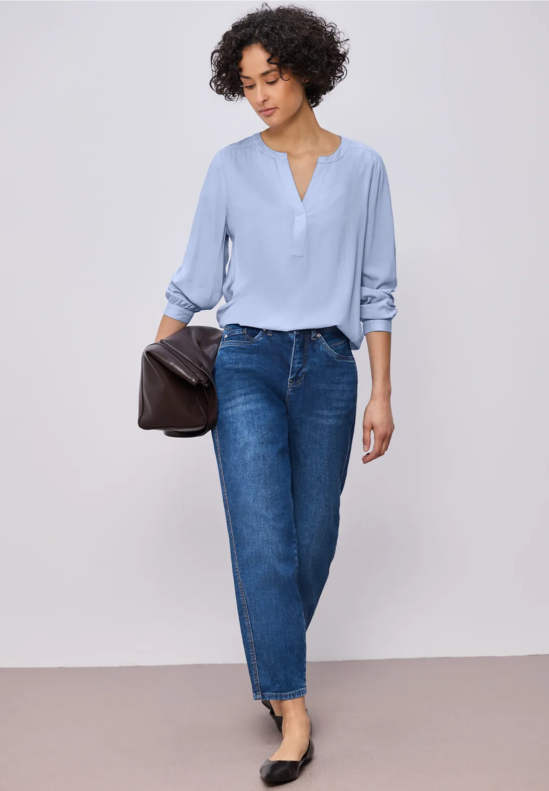Frau in hellblauer Bluse, hoch sitzenden Jeans und schwarzen Schuhen mit dunkelbrauner Handtasche.
