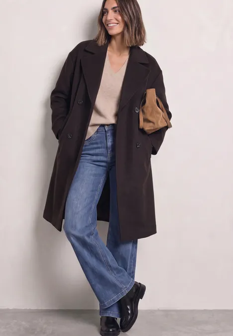 Mantel mit Reverskragen und Wollanteil auburn brown Frau in dunklem Mantel, beigem Pullover, weiten Jeans und schwarzen Stiefeln hält eine Clutch.