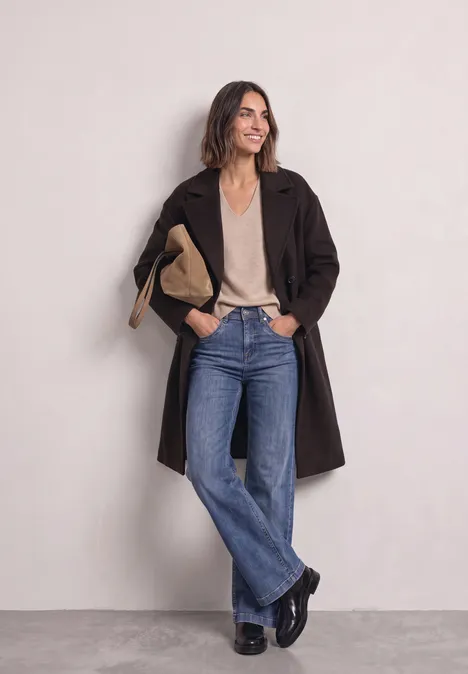 Mantel mit Reverskragen und Wollanteil auburn brown Frau in braunem Mantel, beigem Top, weiten Jeans und schwarzen Schuhen vor hellem Hintergrund.