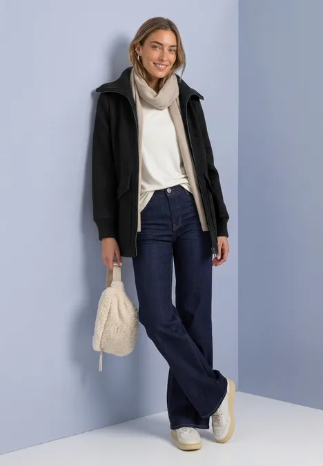 Vrouw in witte trui, sjaal en donker jack, flared jeans, witte sneakers en lichte tas, staand voor een blauwe wand.