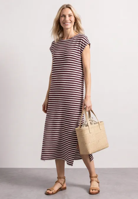 Vrouw in gestreepte midijurk, sandalen en rieten tas voor eenvoudige achtergrond.