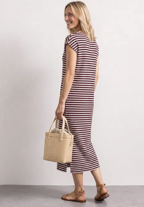 Vrouw in rood-wit gestreepte midijurk met strooien tas en sandalen voor een lichte achtergrond.