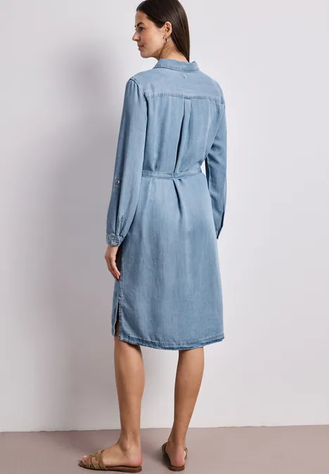 Frau von hinten in einem knielangen Jeanskleid mit langen Ärmeln, barfuß.