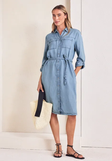 Frau in hellblauem Jeans-Hemdkleid mit Gürtel, trägt Strohtasche und schwarze Sandalen.