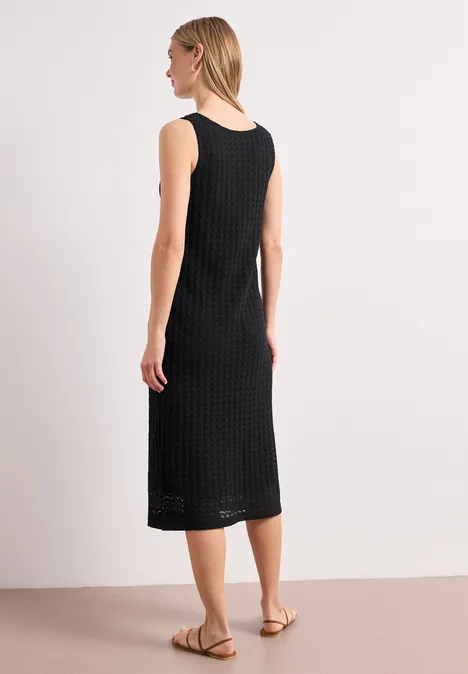 Frau im ärmellosen, schwarzen Midikleid mit Struktur, seitlich stehend, flache Sandalen, heller Hintergrund.