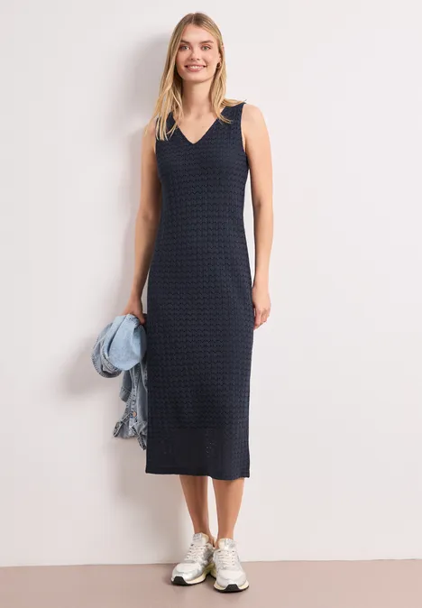 Femme en robe midi sans manches bleu marine à carreaux, baskets blanches et veste en jean posée sur l'épaule.