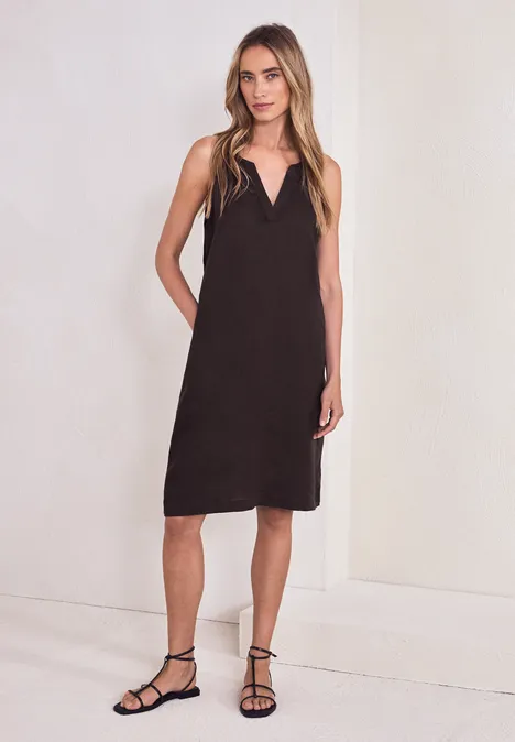 Frau in schlichtem schwarzem ärmellosem Midikleid und flachen Riemchensandalen vor hellem Hintergrund.