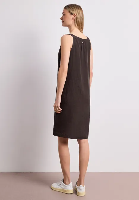 Seitliche Ansicht einer Frau in braunem ärmellosem Kleid und weißen Sneakern.