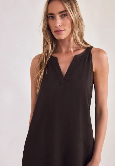 Frau mit langem blonden Haar in ärmellosem schwarzem Kleid mit V-Ausschnitt vor neutralem Hintergrund.