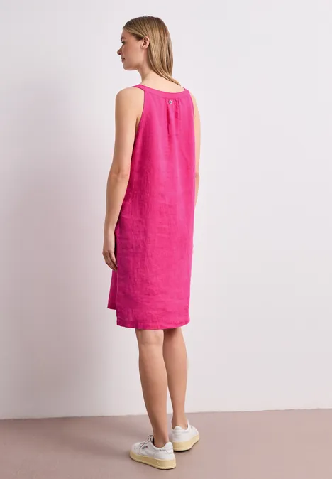 Vrouw van opzij in een knielange, mouwloze linnen jurk fuchsia roze met witte sneakers