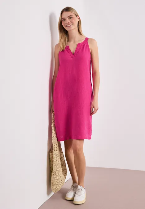 Vrouw in fuchsia mouwloze jurk tot op de knie, witte sneakers en gevlochten tas, leunend tegen een lichte muur.