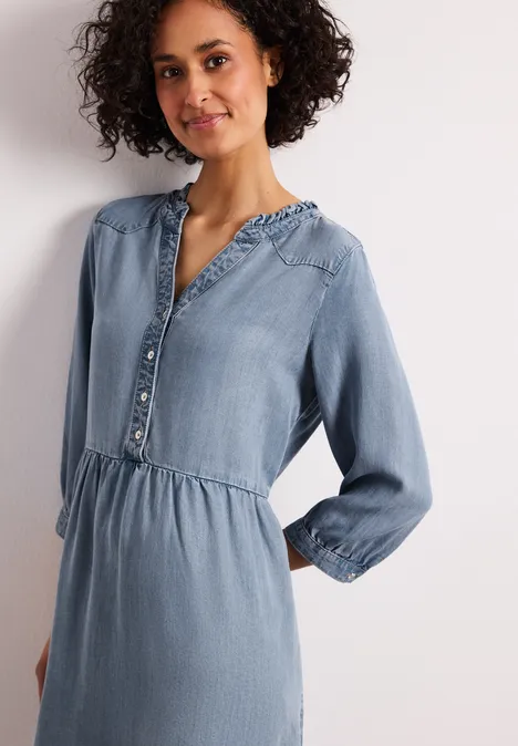 Vrouw in lichtblauwe spijkerjurk met knopen, elastische taille en lange mouwen.