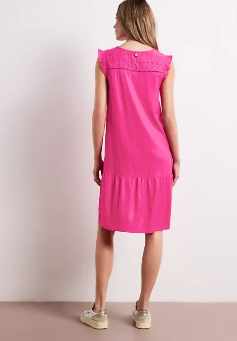 Frau im locker sitzenden pinken, knielangen Kleid mit Spitzenbesatz am Rücken und beigen Sneakern.