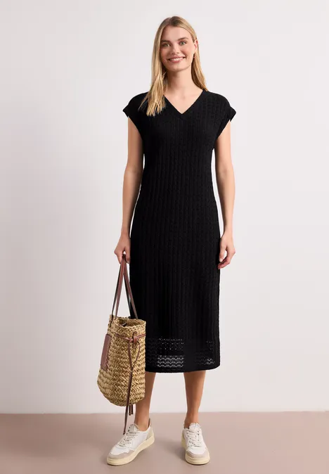 Frau in schwarzem, gestricktem Midikleid mit V-Ausschnitt, weiße Sneaker, hält geflochtene Strohtasche.