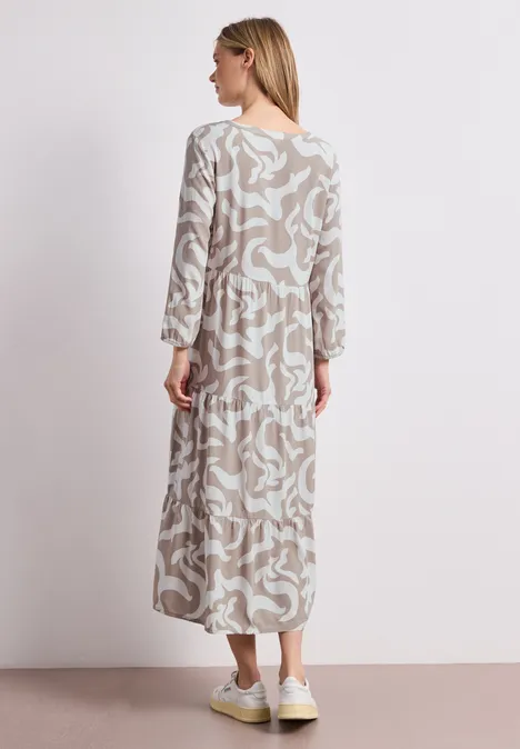 Eine Frau steht mit dem Rücken zur Kamera, trägt ein beiges, weiß gemustertes Maxikleid und weiße Sneaker.