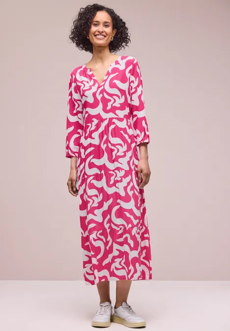 Frau in einem knöchellangen pink-weißen Kleid mit abstraktem Muster und weißen Sneakern.