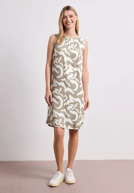 Vrouw in mouwloze, knielange jurk met abstract dessin, witte sneakers, neutrale achtergrond.