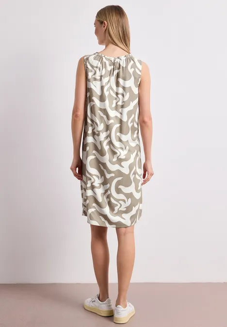 Achteraanzicht van een vrouw in mouwloze jurk met beige abstract dessin en witte sneakers.