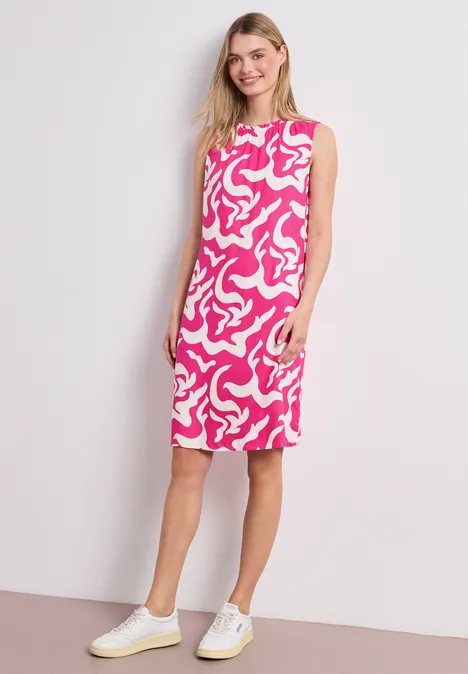 Frau im knielangen, ärmellosen pink-weiß gemusterten Kleid und weißen Sneakern vor neutralem Hintergrund.