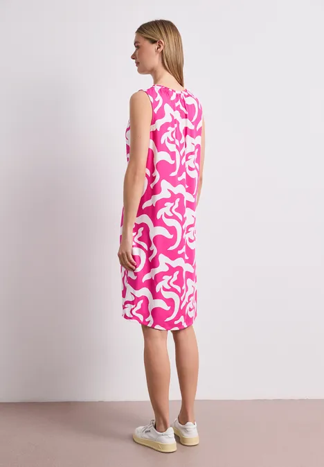 Frau, von hinten, trägt knielanges ärmelloses Kleid mit pink-weißen abstrakten Mustern und weißen Sneakern.