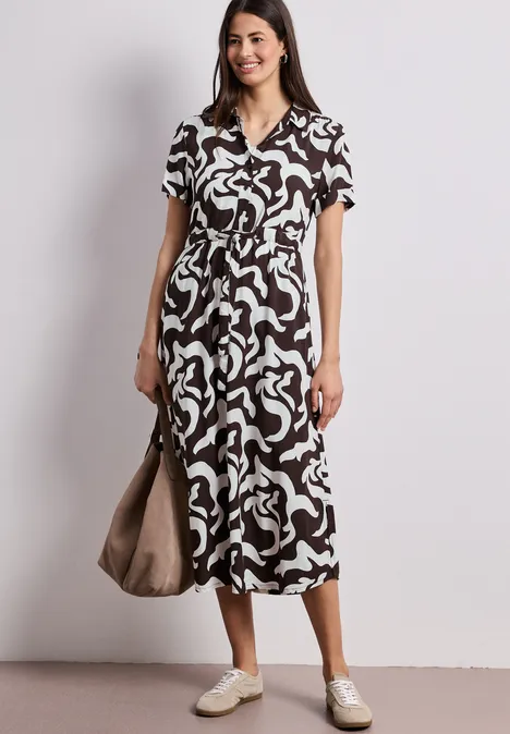 Frau in Midi-Kleid mit abstraktem Schwarz-Weiß-Muster, V-Ausschnitt, weißen Sneakern und Tasche.