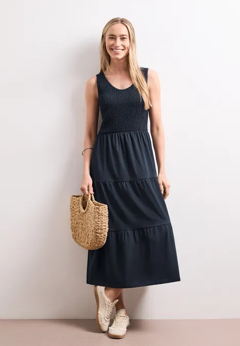 Midi-Kleid mit Smok-Oberteil deep blue Frau trägt dunkelblaues gestuftes Maxikleid, weiße Sneaker und hält Korbtasche.