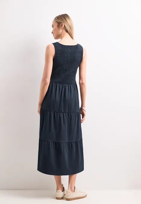 Midi-Kleid mit Smok-Oberteil deep blue Frau von hinten in dunkelblauem, ärmellosem Stufenkleid und hellen Sneakern vor weißem Hintergrund.