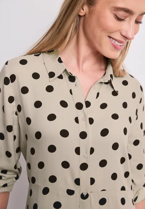 Chemisier beige à pois noirs, col et patte de boutonnage, manches retroussées de façon décontractée.
