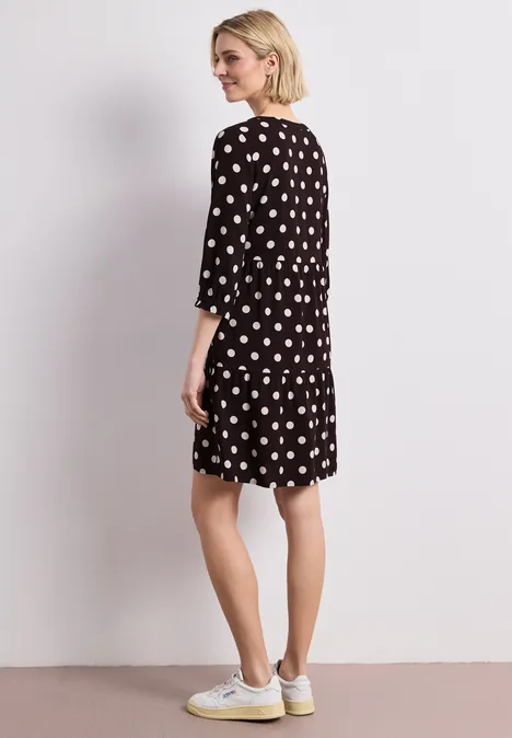 Frau in schwarzem, kurzem Tupfenkleid mit Halblangsärmeln und hellen Sneakern, seitlich stehend vor neutralem Hintergrund.