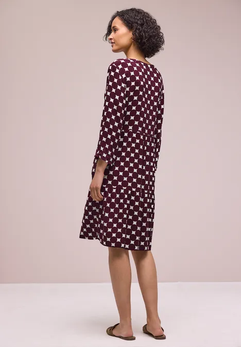 Eine Person mit lockigen Haaren steht in einem weinroten Kleid mit weißen Punkten und Sandalen vor beigem Hintergrund.